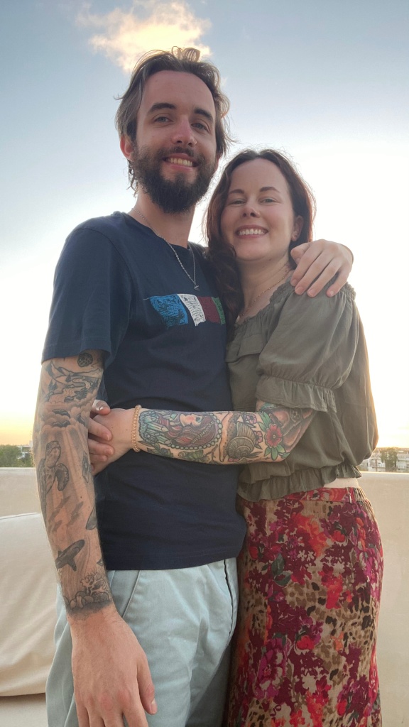 man and woman with arms around each other smiling in Mexico on a rooftop with a sunset behind them