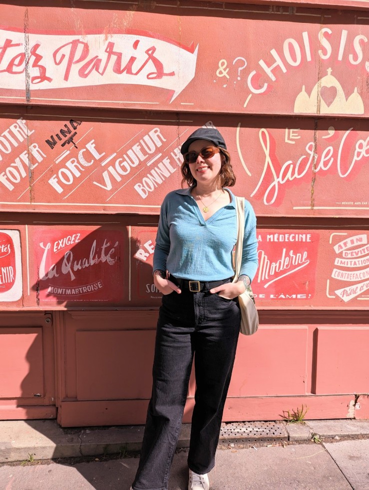 woman standing in front of a red wall with French words written all over it in Paris France.  She is wearing a collared blue sweater tucked into black wide leg jeans.  she is carrying a totebag and wearing a grey newsboy hat and white sneakers.
